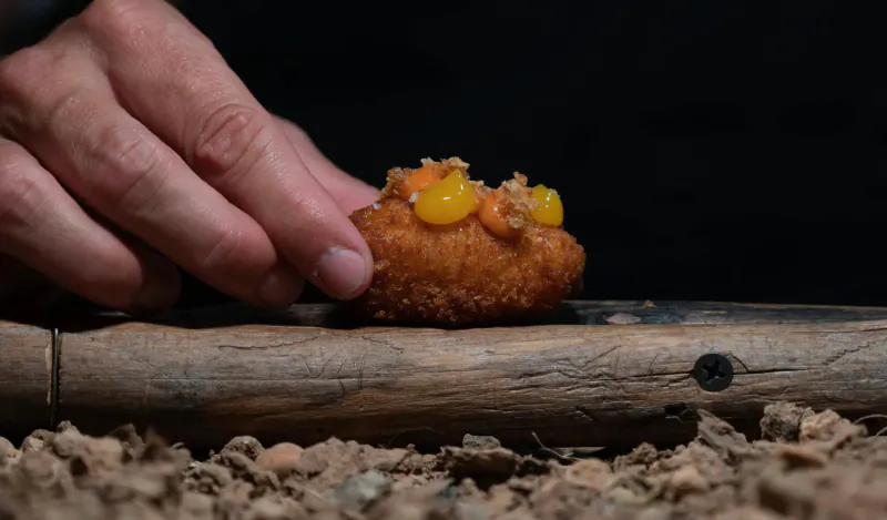Hand gently touching crispy golden croquette topped with orange roe on wooden board at Michelin-starred restaurant.