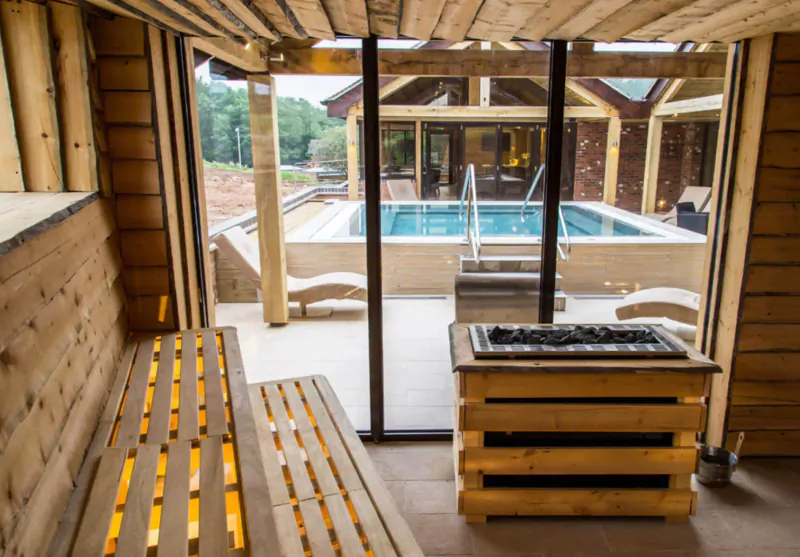 Wooden sauna interior with tiered benches, fire pit, and view of outdoor infinity pool at Moddershall Oaks Spa.