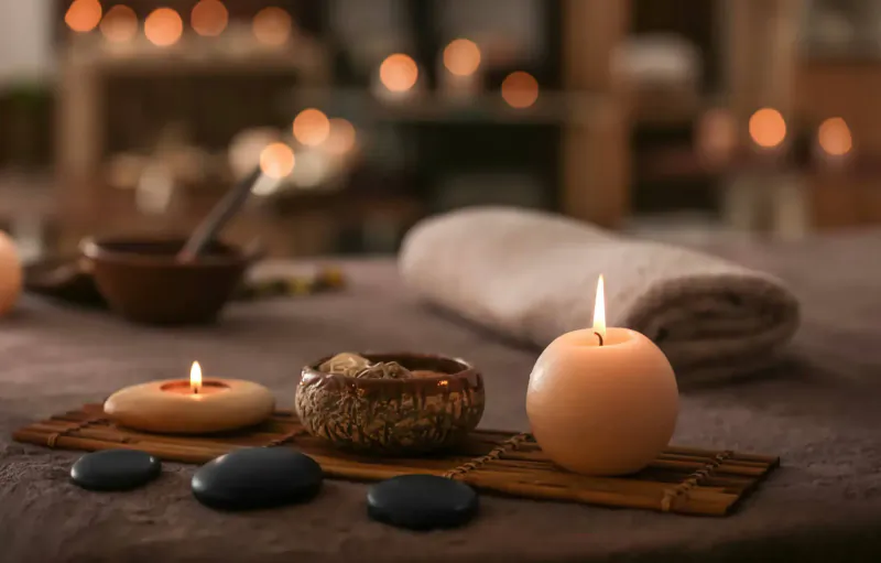 Spa table with lit candles, black stones, ceramic bowls, and folded towel in warm ambient light.
