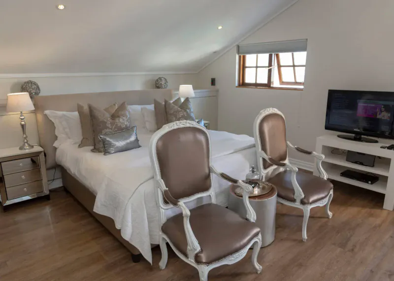 Luxurious attic bedroom at Turbine Boutique Hotel with white king bed, beige velvet chairs, lamps, and TV.