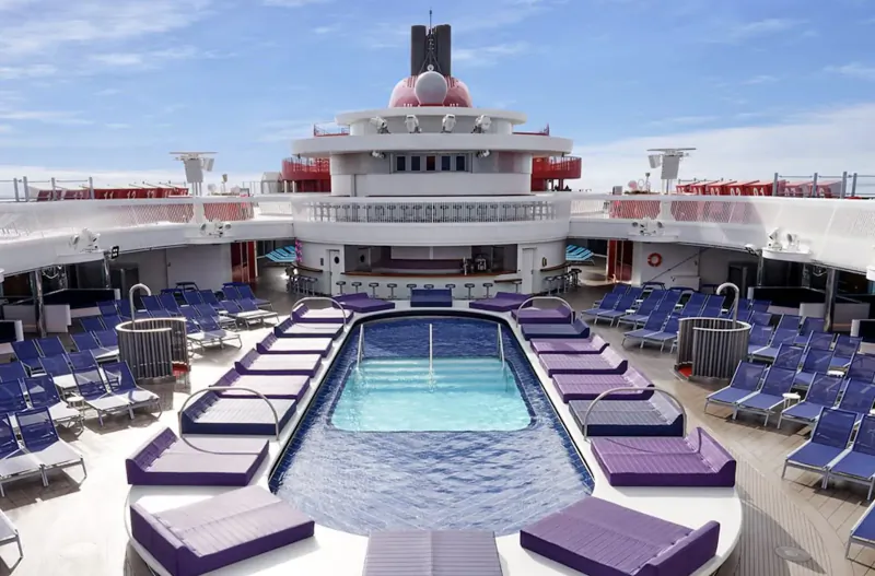 Virgin Voyages cruise ship deck with rectangular pool, purple loungers, blue chairs, and sunny sky.