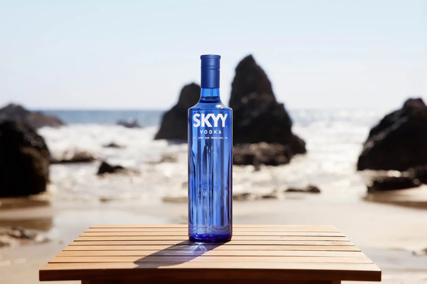 Blue SKYY Vodka bottle on wooden table at beach with ocean waves and rocks, sunny sky.
