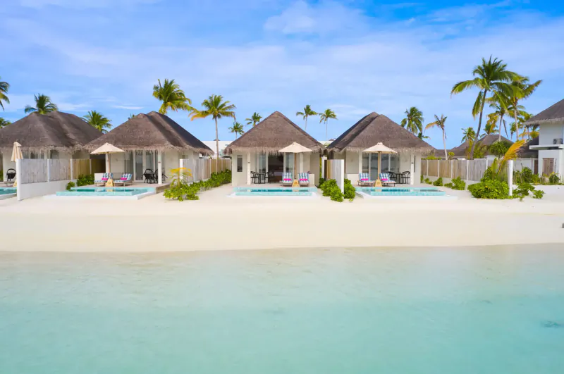 Luxury thatched beachfront villas with private pools on white sand beach and turquoise lagoon, surrounded by palm trees.