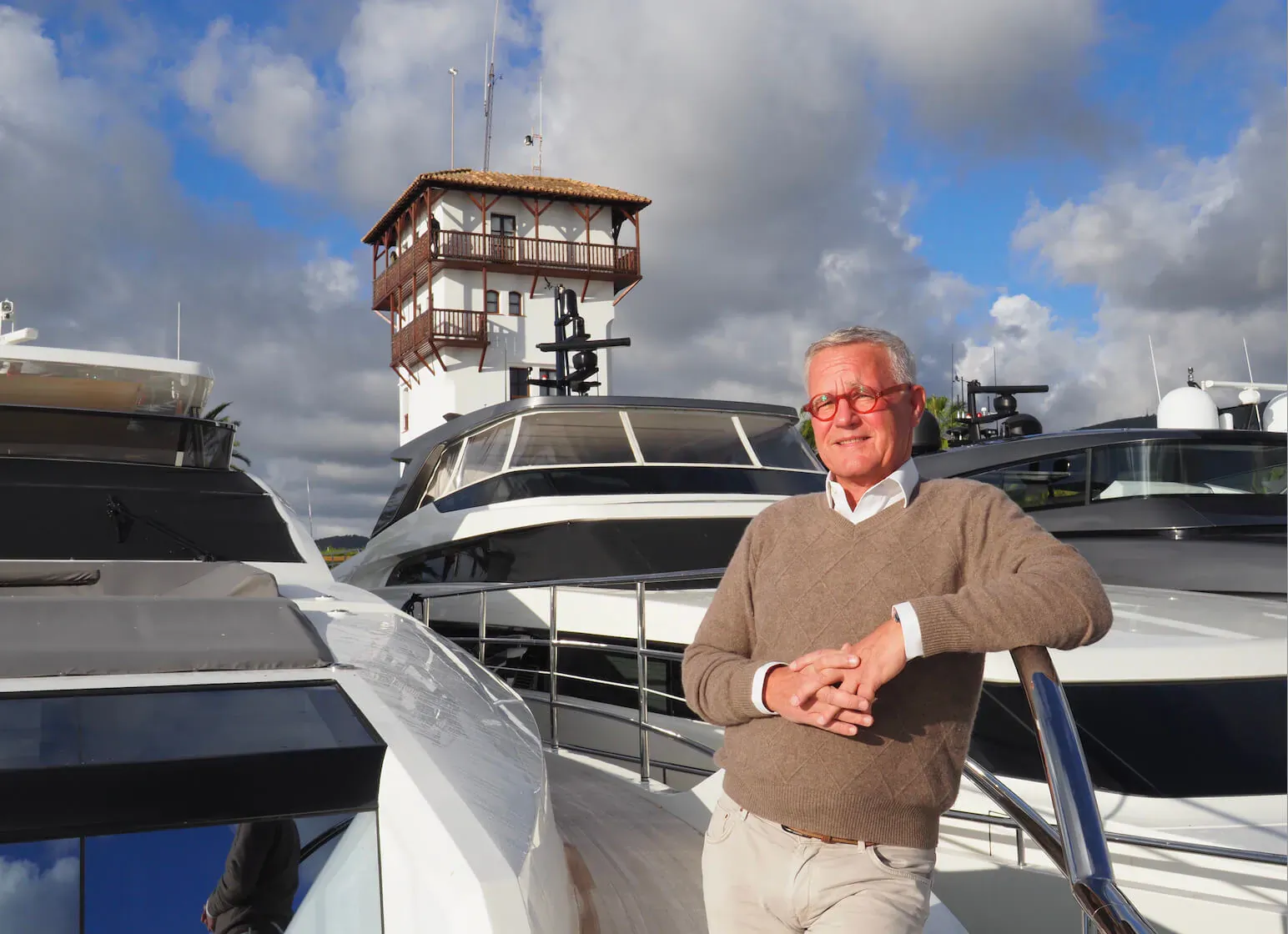 Andreas Hoffmann in beige sweater leaning on white yacht railing, marina with tower and cloudy sky behind.