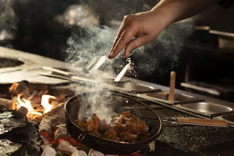 Chef's hand sprinkling seasoning on sizzling fried chicken in hot wok over flames at Chotto Matte Soho kitchen