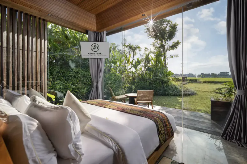 Spacious villa bedroom at Gdas Bali Resort with 'Gdas Bed' sign, king bed, chair, table, and lush rice field view through glass walls.