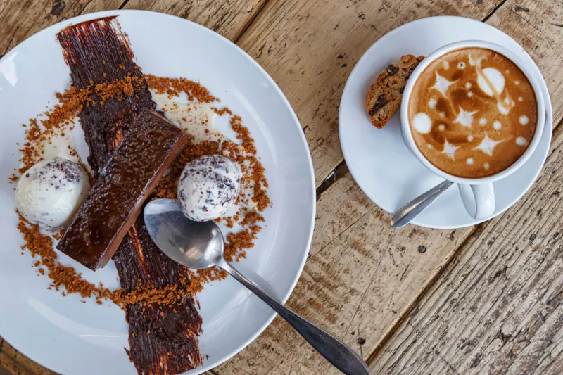Chocolate brownie with ice cream scoops and bacon strip on rustic wooden table beside cappuccino with foam art