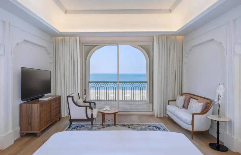 Luxurious hotel suite with white bed, ornate furniture, TV, and arched window overlooking sea balcony.