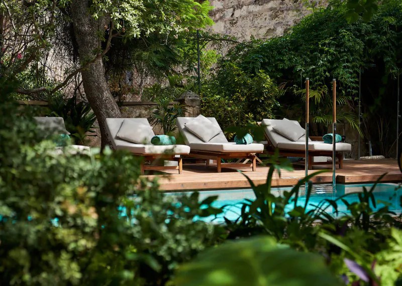 Row of cushioned lounge chairs with green towels by turquoise pool in lush garden at Can Bordoy Grand House & Garden