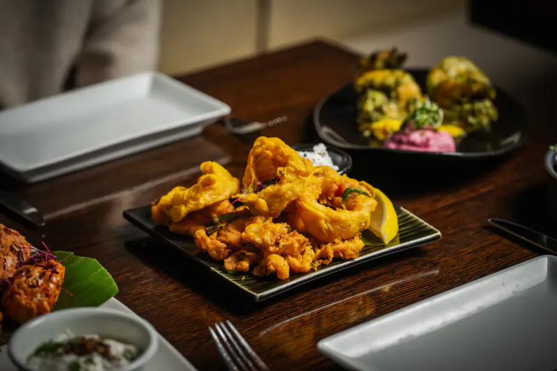 Fried calamari rings on black square plate with lemon wedge and dip, on wooden table with Indian dishes nearby