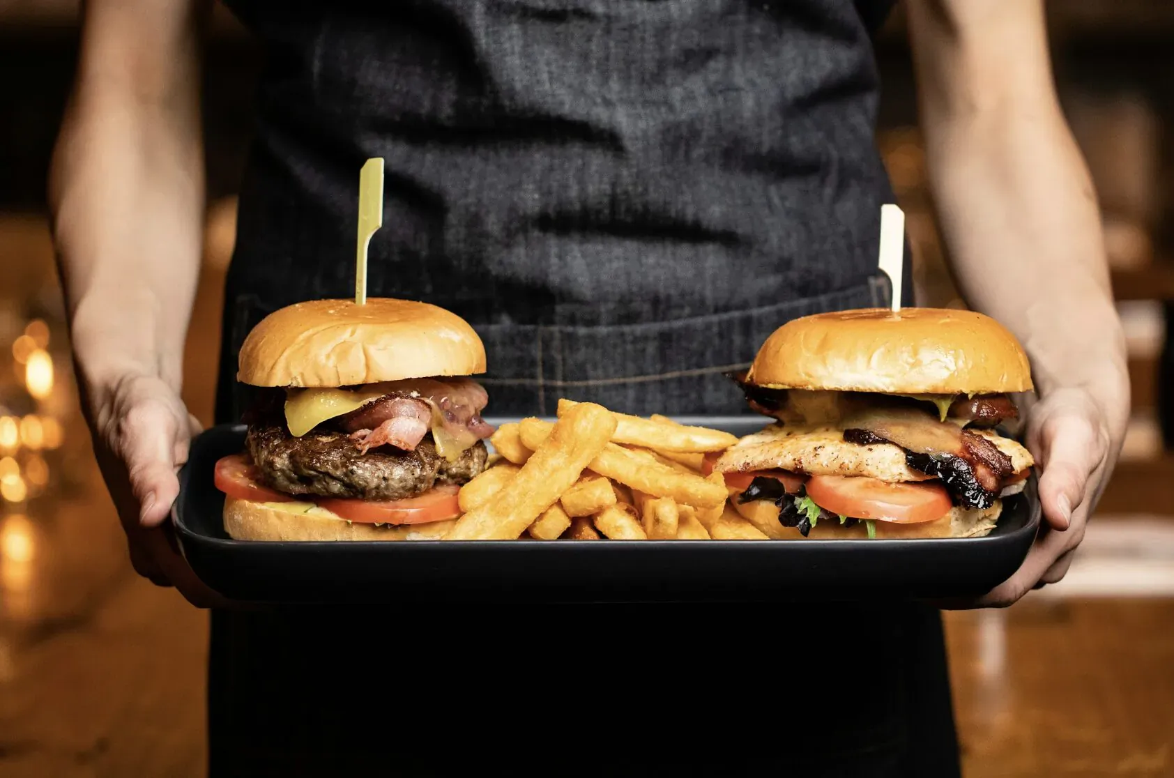 Server holding tray with two garnished burgers and fries in restaurant setting