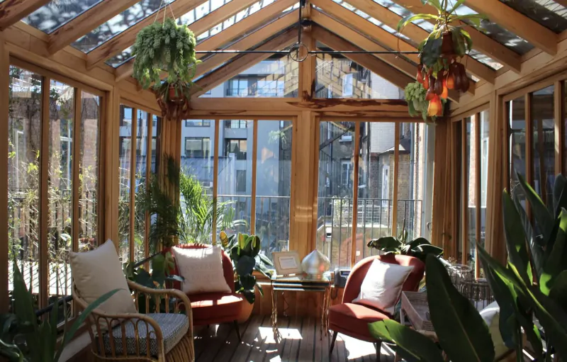 Sunlit wooden conservatory with hanging plants, lush greenery, white and red cushioned chairs around glass table