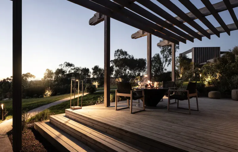 Twilight view of wooden deck at ADLER Spa Resort Sicily with fire table, chairs, lush gardens, and path.