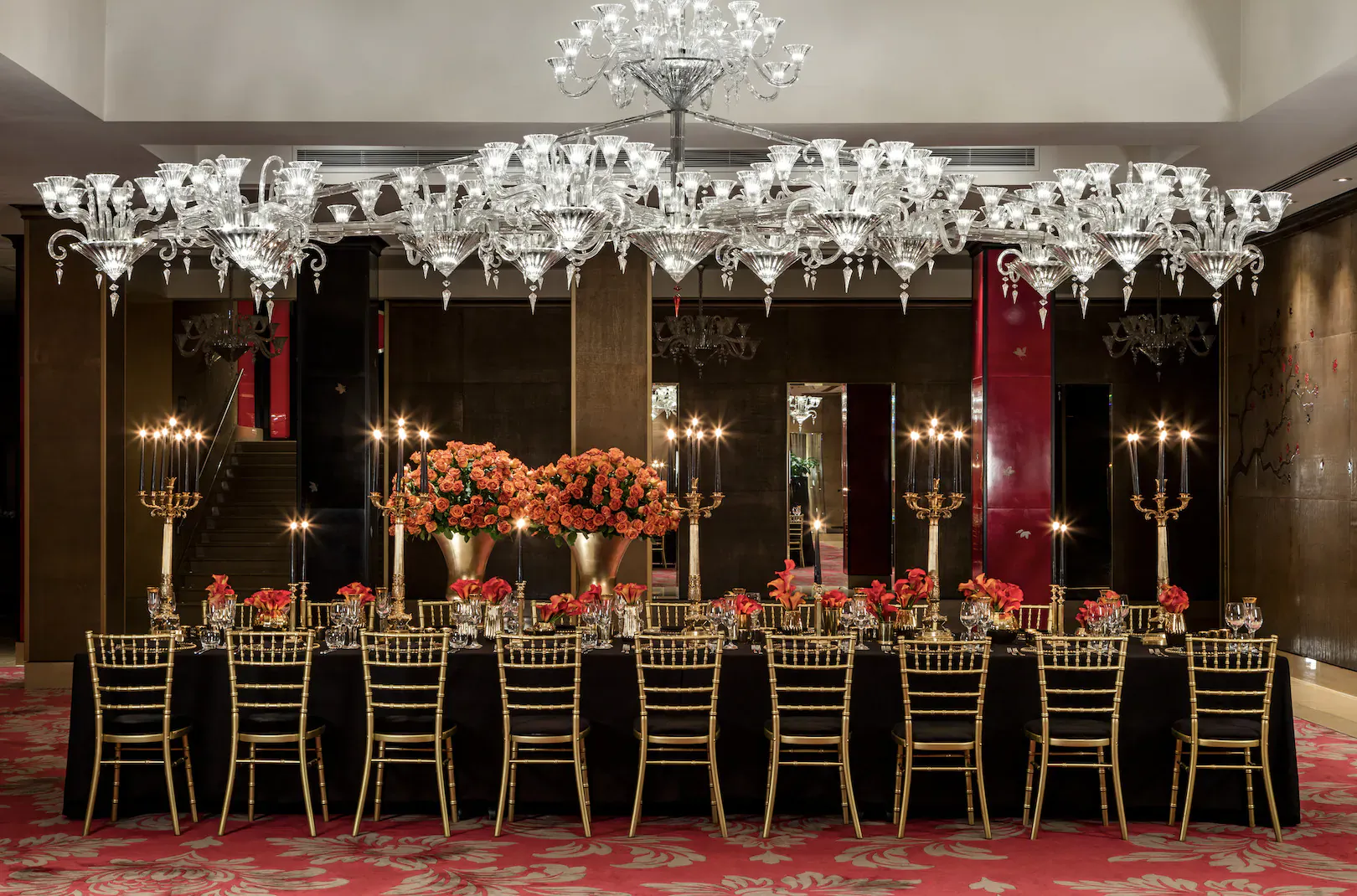 Elegant hotel dining room with crystal chandeliers, red walls, orange floral centerpieces on long black table with gold chairs