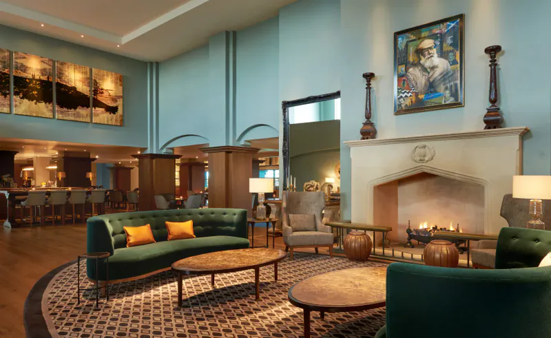 Elegant hotel lounge with green velvet sofas, wooden coffee table, lit fireplace, abstract art, and bar area.