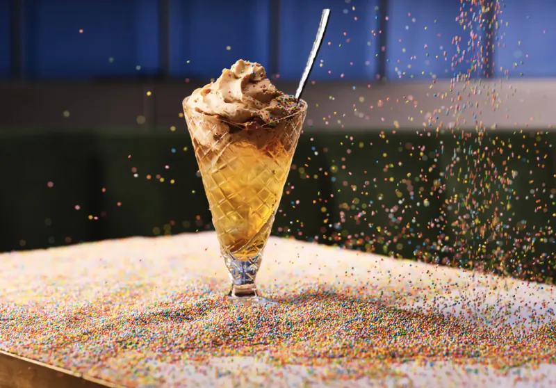 Faceted glass of amber cocktail topped with cream and spoon, surrounded by falling colorful sprinkles on table at rooftop bar