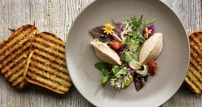 Grilled chicken salad with greens, edible flowers, and tomatoes on a plate, flanked by toasted bread slices on wooden surface