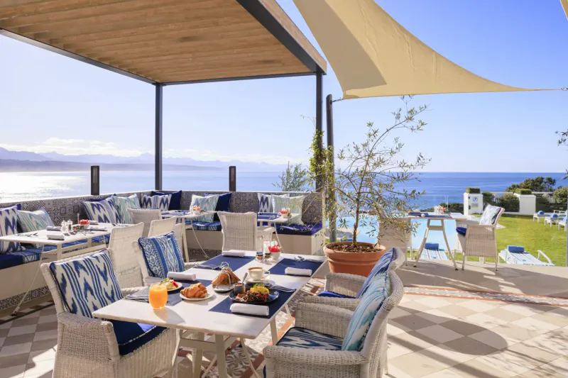 Outdoor terrace breakfast at The Plettenberg Boutique Hotel with blue-cushioned chairs, croissants, orange juice, and ocean view.