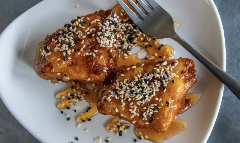 Two crispy sesame chicken pieces glazed with honey sauce on a white plate, pierced by a fork.