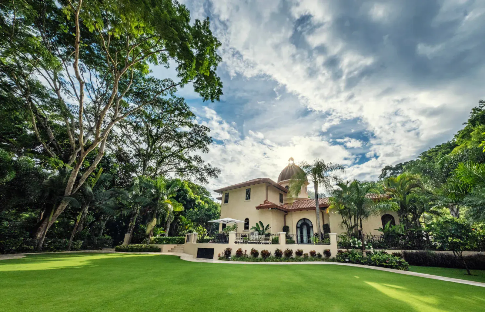 Beige villa with red tile roof and dome, surrounded by palm trees and lush green lawn under partly cloudy sky