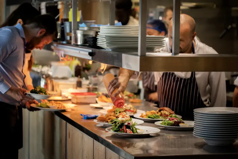 Busy kitchen staff plating salads, pouring sauce, and stacking dishes on counter under warm lights