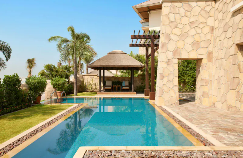 Luxury villa poolside at Sofitel Dubai the Palm with turquoise infinity pool, thatched pergola, palms, stone walls, lush gardens under blue sky.