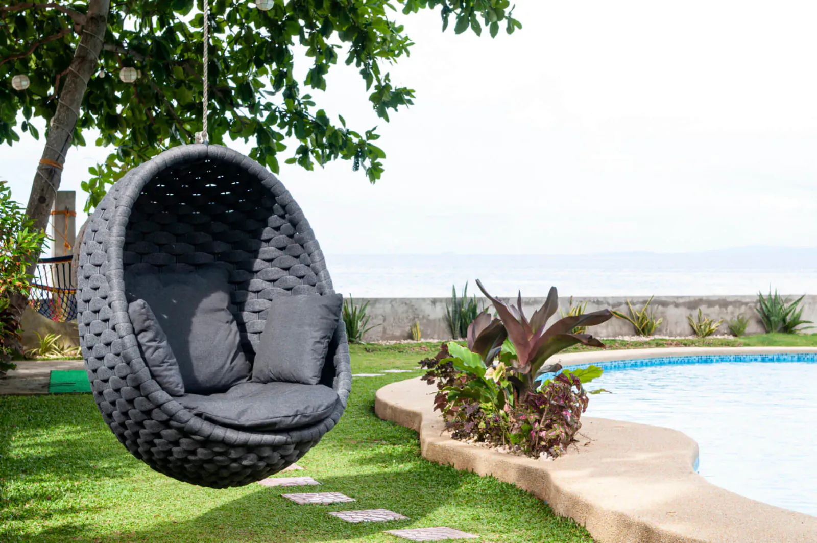 Gray woven egg-shaped hanging chair suspended from tree near tropical plants, pool, and garden in resort setting.
