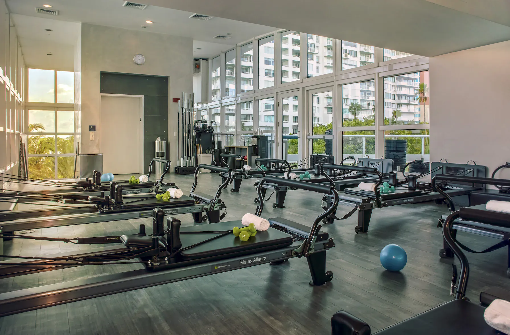 Spacious modern Pilates studio with reformer machines, exercise balls, towels, and large windows overlooking cityscape.