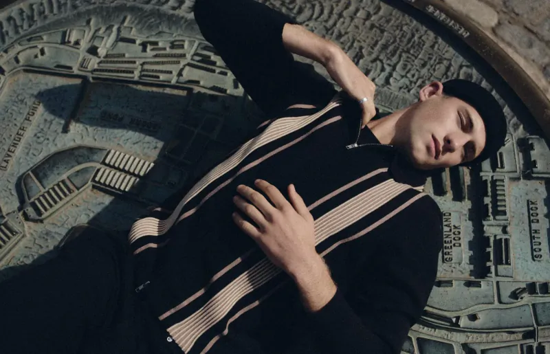 Young man in black and white striped cardigan and beanie lies reclined on detailed cityscape sculpture in Canary Wharf.