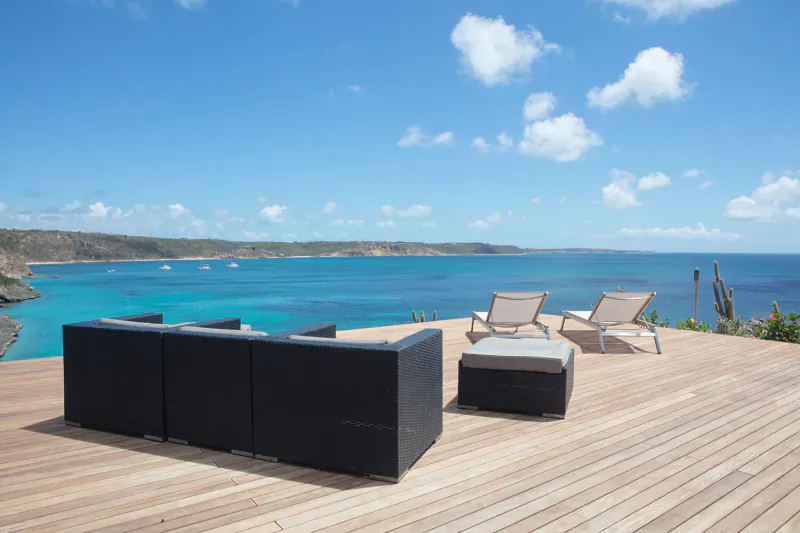 Modern black outdoor sofa set and loungers on wooden deck overlooking turquoise sea and cliffs at private resort