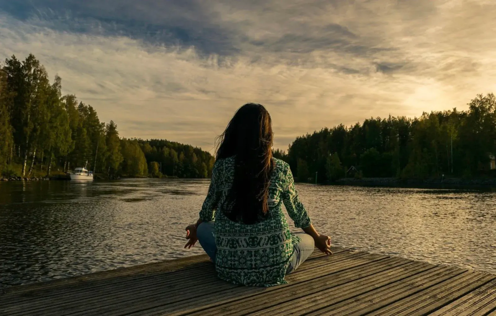 Woman in green tunic sits in lotus pose on dock, meditating by lake at sunset with boat and forest.