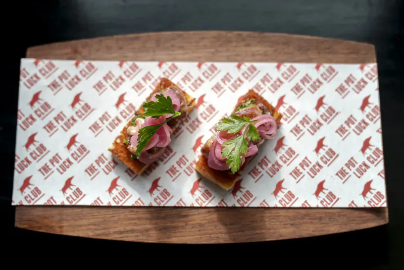 Two The Pot Luck Club open-faced sandwiches with pink pickled onions and herbs on a wooden board with branded paper.