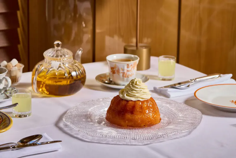Orange dome pudding topped with cream on glass plate, with teapot, cups, lemon water on white tablecloth at Italian ristorante.