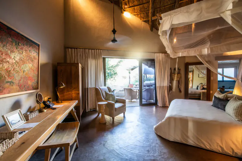 Spacious Tarkuni suite at Tswalu with king bed under mosquito net, wooden desk, chairs, and sunset view through open doors.