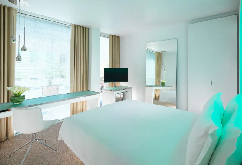 Modern hotel room at St. Martins Lane with white bed, teal accents, desk, TV, and city view through large windows.