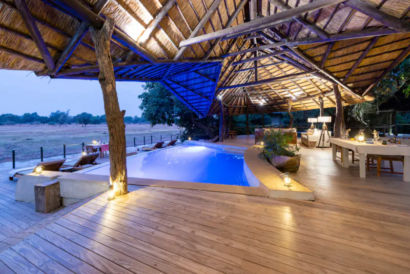 Twilight view of Lion Camp's elevated wooden deck with glowing blue infinity pool, lounge chairs, tables, and savanna backdrop.