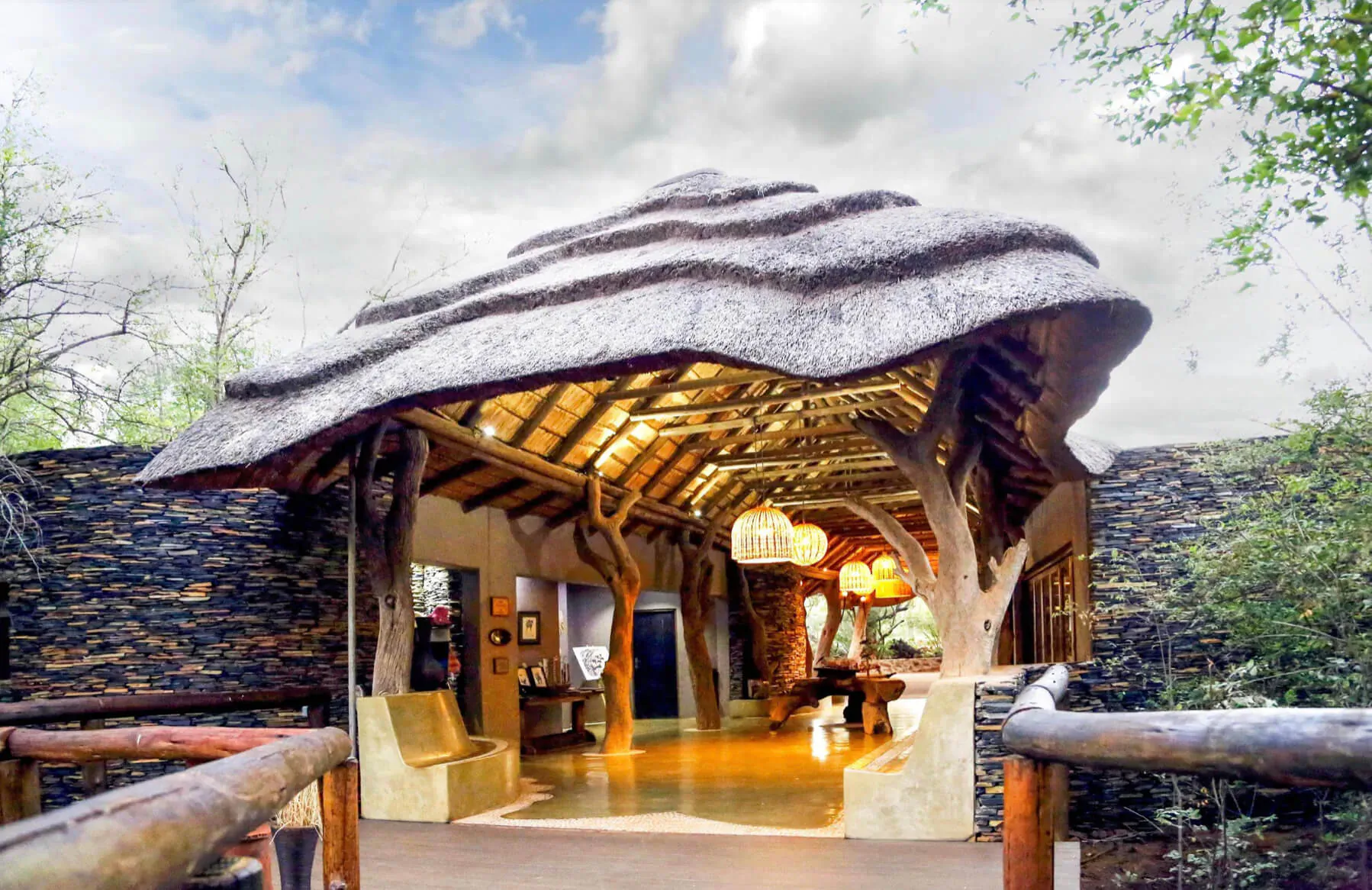 Madikwe Hills Private Game Lodge entrance: thatched roof structure with wooden beams, stone walls, lush greenery, and open lounge area.