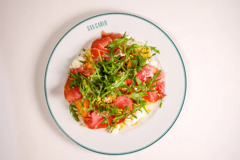 White plate with 'San Carlo' logo, topped with sliced tomatoes, arugula, and white cheese on a white surface