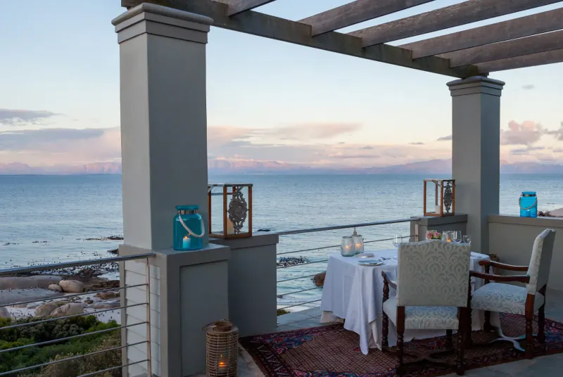 Elegant outdoor dining table set for two on a terrace overlooking the ocean at sunset, with lanterns and columns at Tintswalo Boulders.