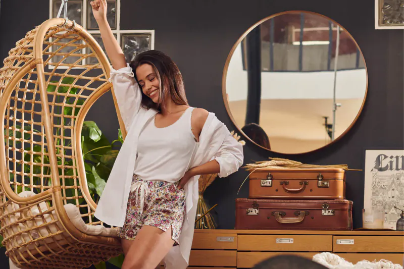 Smiling woman in white tank top, printed shorts, open kimono sits in rattan hanging chair, beside gold mirror, vintage suitcases, and Le Colonel poster
