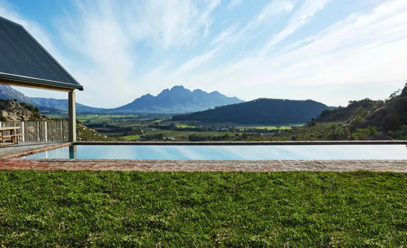 Infinity pool at modern house with pergola overlooking Franschhoek mountains and valley under blue sky.