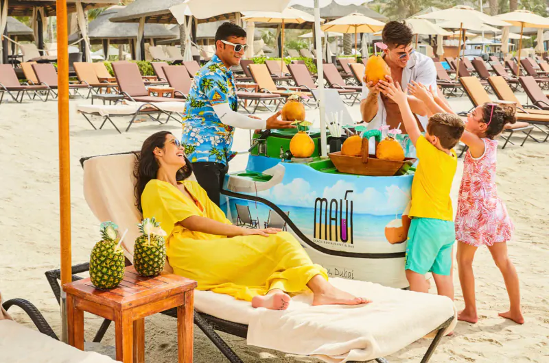 Family and staff at Mafr fruit stand on Dubai beach, serving fresh oranges and pineapples by pool loungers.