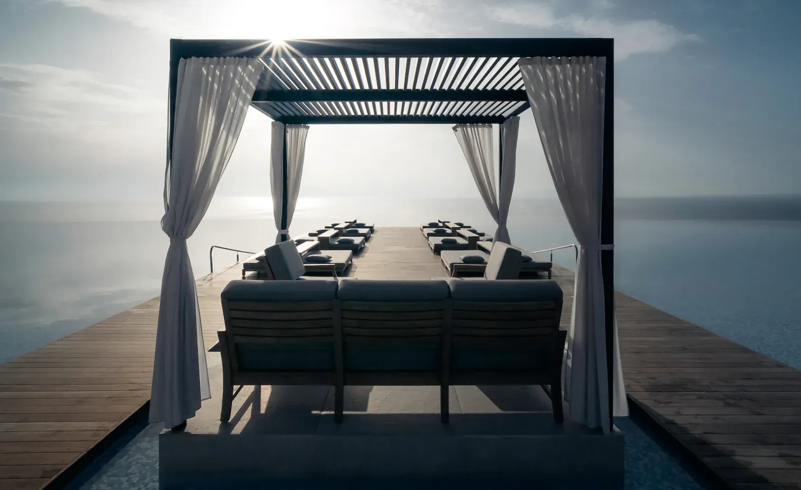 Luxury open-air cabana with long dining table, cushioned bench, and sheer curtains on a wooden pier over calm sea at sunset