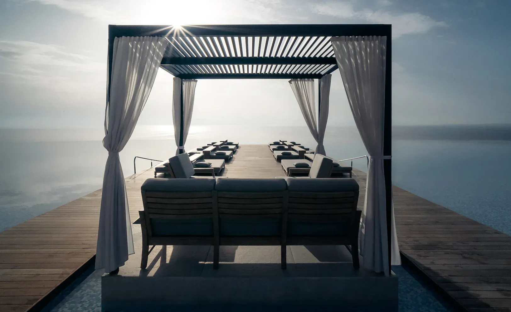 Luxury open-air cabana with long dining table, cushioned bench, and sheer curtains on a wooden pier over calm sea at sunset