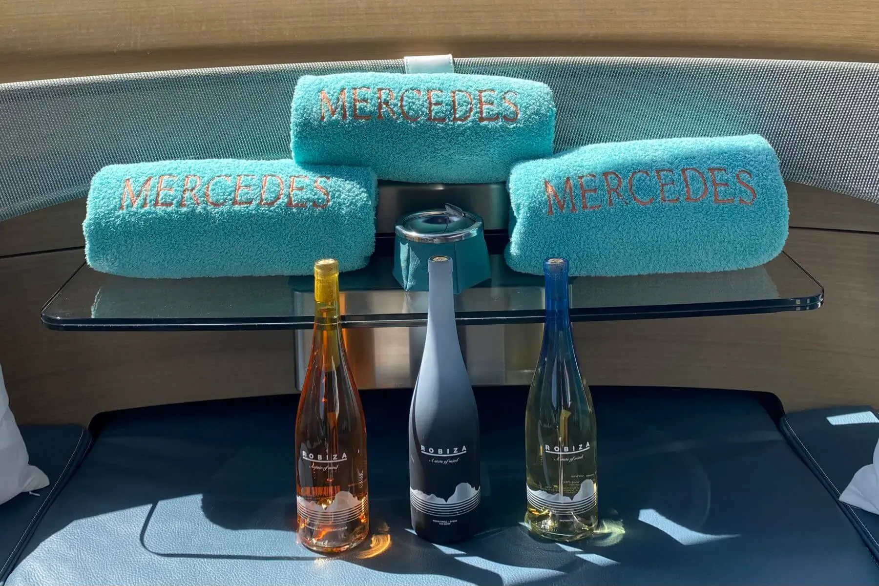 Mercedes-branded teal towels and Robiza rosé, white, and gray wine bottles on glass table in luxury yacht lounge.