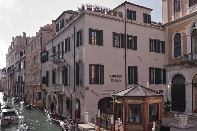 Violino d'Oro Hotel exterior in Venice canal-side, with shuttered windows, plants on roof, and entrance kiosk.