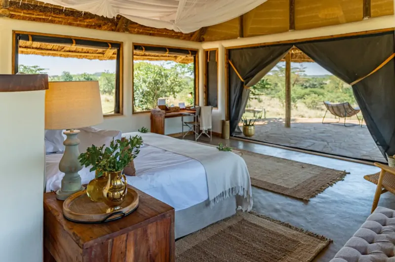 Spacious safari lodge bedroom with king bed, wooden nightstand, lamp, and open doors to savanna view.