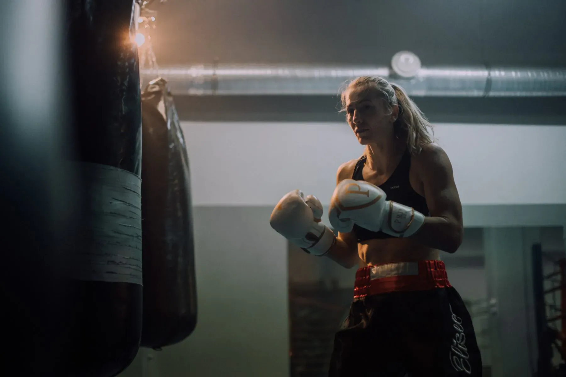 Blonde woman in boxing gloves and gear punching a heavy bag in a dimly lit gym studio.