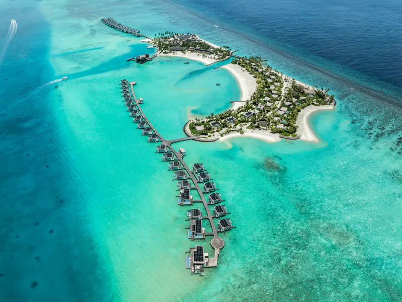 Aerial view of Crossroads Maldives resort: overwater villas on turquoise lagoon surrounding lush island, sustainable living.