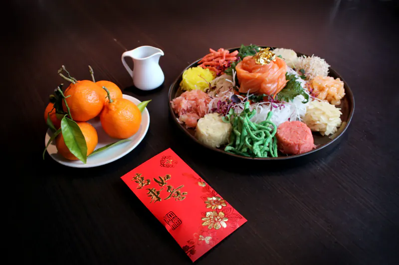 Luxurious yu sheng salad with salmon rose, rice, veggies on dark table, flanked by oranges, sauce jug, red packet.